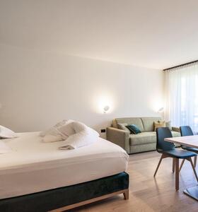 Doppelzimmer mit Doppelbett, Sofa und Sitzbereich mit Holztisch und gepolsterten Stühlen vor einer großen Fensterfront.