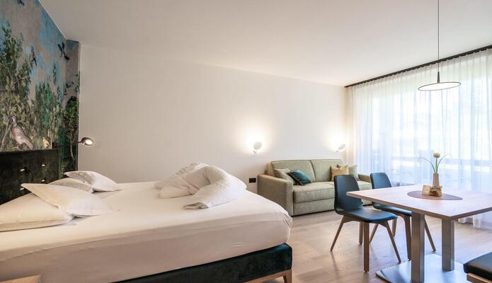 Doppelzimmer mit Doppelbett, Sofa und Sitzbereich mit Holztisch und gepolsterten Stühlen vor einer großen Fensterfront.