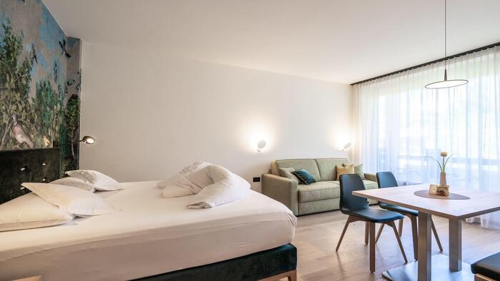 Camera doppia con letto matrimoniale, divano e zona salotto con tavolo in legno e sedie imbottite davanti a un’ampia vetrata.