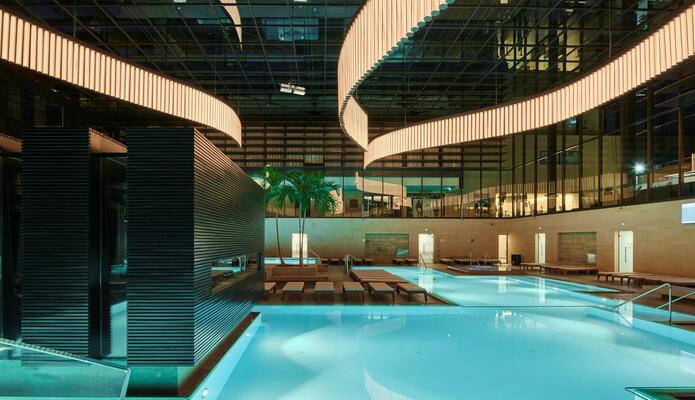 Terme Merano con piscine illuminate, lettini, palme e una grande installazione luminosa sul soffitto.