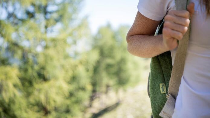 Primo piano di una persona che cammina con uno zaino verde, con alberi sfocati sullo sfondo.