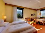 Rosenapfel double room with a double bed, seating area with a wooden table and red upholstered seating, and large windows overlooking greenery.