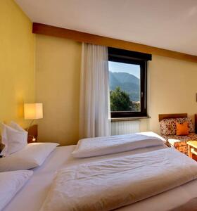 Doppelzimmer Rosenapfel mit Doppelbett, Sitzbereich mit Holztisch und roten Polstermöbeln sowie großen Fenstern mit Blick ins Grüne.