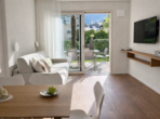 Heller Wohnbereich mit Sofa, Couchtisch, TV-Wand, Esstisch und Zugang zum Balkon mit Blick auf die Landschaft.