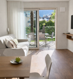 Zona giorno luminosa con divano, tavolino, parete TV, tavolo da pranzo e accesso al balcone con vista sul paesaggio.