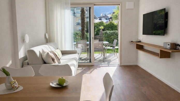 Zona giorno luminosa con divano, tavolino, parete TV, tavolo da pranzo e accesso al balcone con vista sul paesaggio.