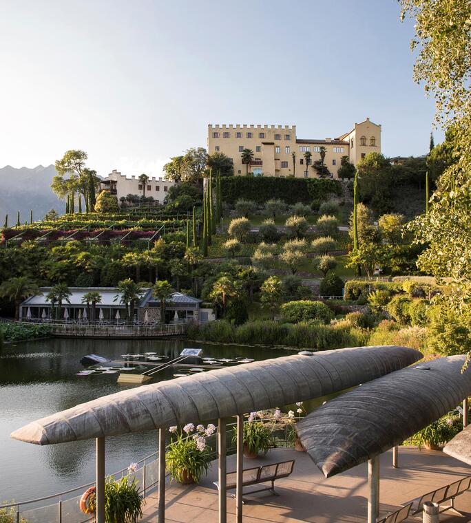 Vista su un laghetto e un’area con panchine coperte, con i giardini terrazzati e Castel Trauttmansdorff sullo sfondo.