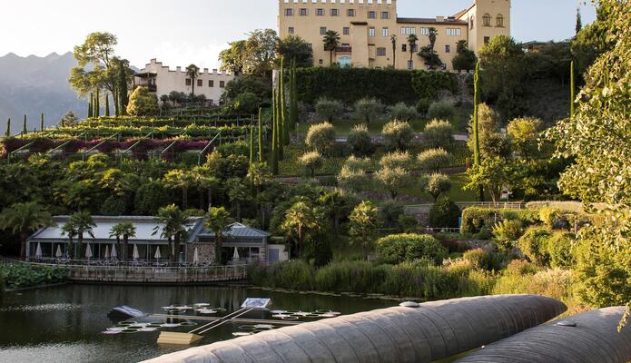 Blick über einen Teich und überdachte Sitzplätze auf die terrassenförmigen Gärten und Schloss Trauttmansdorff.