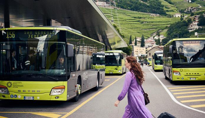 Frau im violetten Kleid geht mit einem Rollkoffer über einen Zebrastreifen im Hintergrund sind Busse.