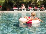 A woman relaxes on an inflatable ring in the outdoor pool, while a man sits on a red sun lounger in the lush garden.