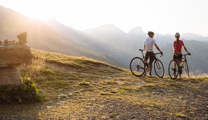 Due ciclisti su un sentiero di montagna, con montagne sullo sfondo e prati soleggiati in primo piano.