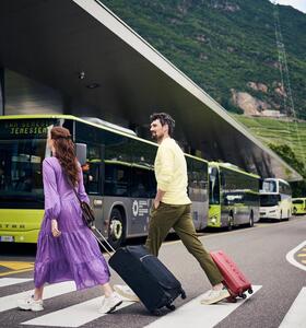 Alcune persone con valigie con le ruote attraversano un passaggio pedonale, con autobus parcheggiati sullo sfondo.