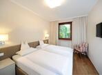 Zimmer im Apartment Palmengarten mit Doppelbett, Fernseher an der Wand, einem Sessel neben dem Fenster mit Blick ins Grüne.