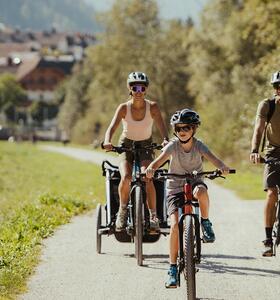 Ciclista su un ampio sentiero tra prati e alberi, con case sullo sfondo.